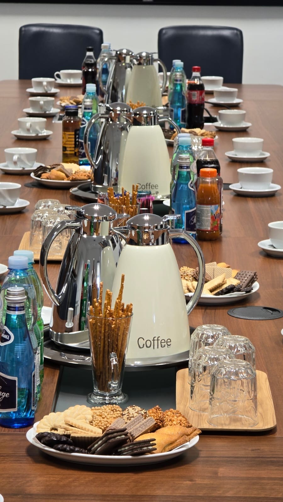 Długi stół konferencyjny z ciemnego drewna, nakryty białymi filiżankami, spodkami, talerzykami z ciastkami, słonymi paluszkami w szklance oraz termosami do kawy i butelkami wody mineralnej oraz soków.