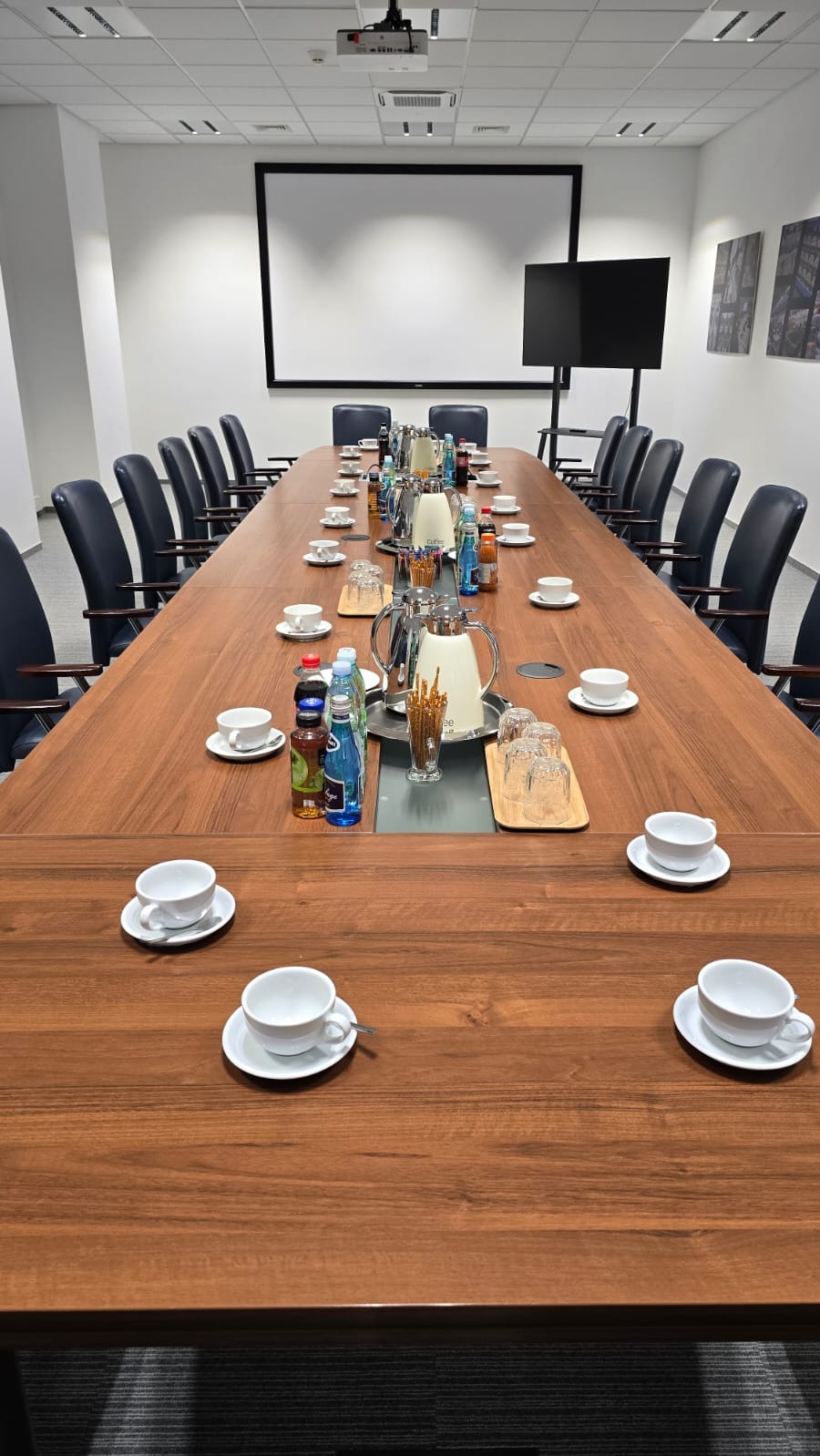 Długi stół konferencyjny z drewnianym blatem, nakryty białymi filiżankami, dzbankami termicznymi, butelkami wody i słonymi paluszkami, przygotowany na spotkanie w jasnym biurze z projektorem...