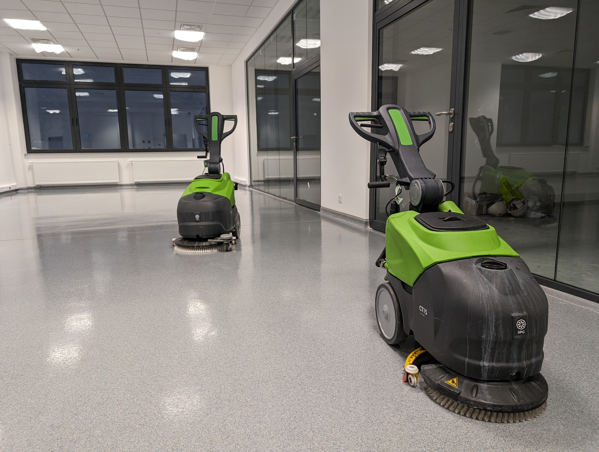 Dwie szorowarki automatyczne, czarno-zielone, stoją na jasnej, wzorzystej podłodze w przestronnym, nowoczesnym biurze z dużymi oknami i szklanymi ścianami działowymi.