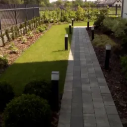 Ułożona ścieżka z szarej kostki brukowej prowadząca przez zadbany ogród z trawnikiem, niskimi krzewami i nowoczesnymi lampami ogrodowymi, widok w kierunku domu z ciemnym ogrodzeniem panelowym.