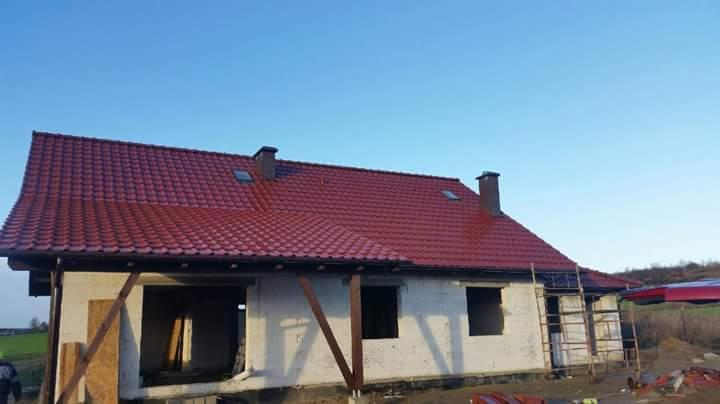 Czerwony dach z dachówki ceramicznej na surowym budynku, widoczne kominy i okna dachowe, w tle błękitne niebo i zieleń, częściowo widoczna drabina.