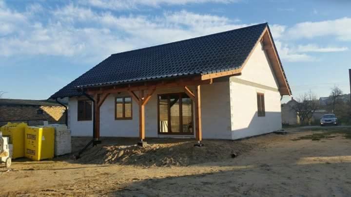 Dom jednorodzinny z ciemną dachówką karpiówką i drewnianą konstrukcją werandy. Widoczne rynny. W tle błękitne niebo z chmurami.