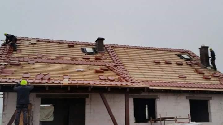 Montaż dachówki na nowym domu z oknami dachowymi i kominem. Dekarze w kaskach układają czerwone dachówki karpiówki na drewnianej konstrukcji dachu. Szary, pochmurny dzień.