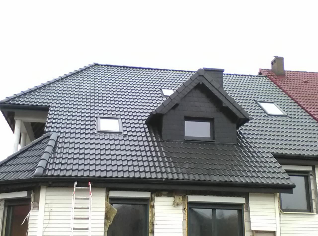 Dom z nowym, ciemnoszarym dachem z dachówki karpiówki i oknami dachowymi. Część elewacji w trakcie wykończenia, widoczna drabina oparta o ścianę.