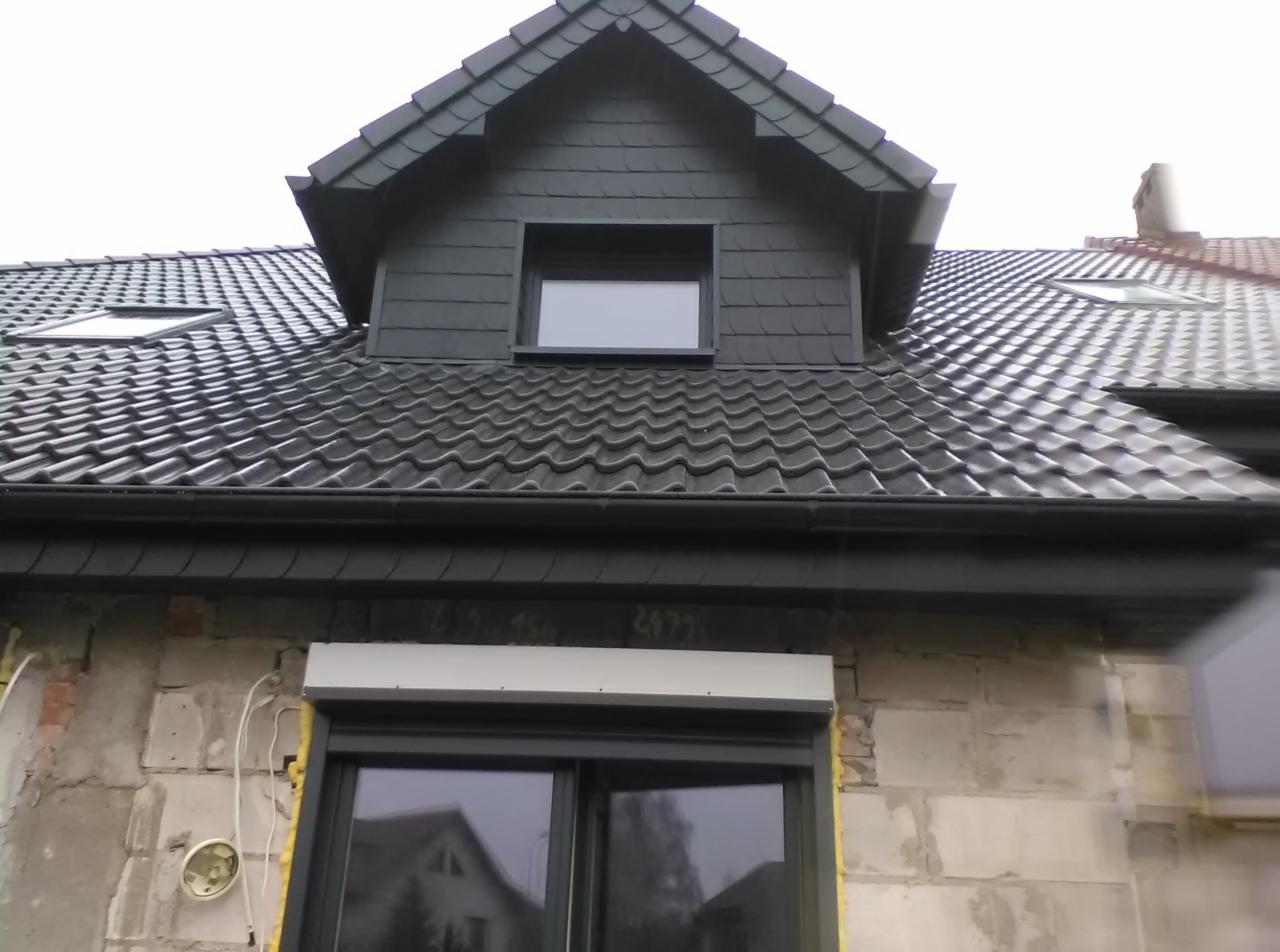 Czarny dach z dachówką karpiówką i lukarną z szarej blachy. Widoczne okna dachowe i rynna. Mur z pustaków. Okno z roletą zewnętrzną.