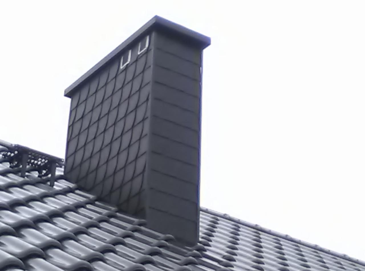 Szary komin obłożony łupkiem na dachu z ciemnej dachówki ceramicznej. Widok z dołu, pochmurne niebo w tle. Fragment barierek kominiarskich.