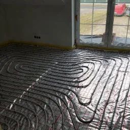 Ułożona instalacja ogrzewania podłogowego na folii izolacyjnej w jasnym pomieszczeniu, widok na okno z dwoma białymi dostawczymi autami na zewnątrz.