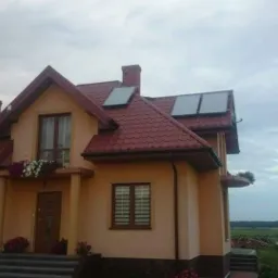 Dom jednorodzinny z panelami słonecznymi na dachu z czerwonej dachówki, kwiatami w oknach i widokiem na okolicę w tle.