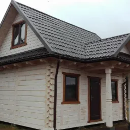 Nieduży, parterowy budynek z bali, z ciemnym dachem dwuspadowym i drewnianą stolarką okienną, widoczny z boku, na tle zalesionego terenu.