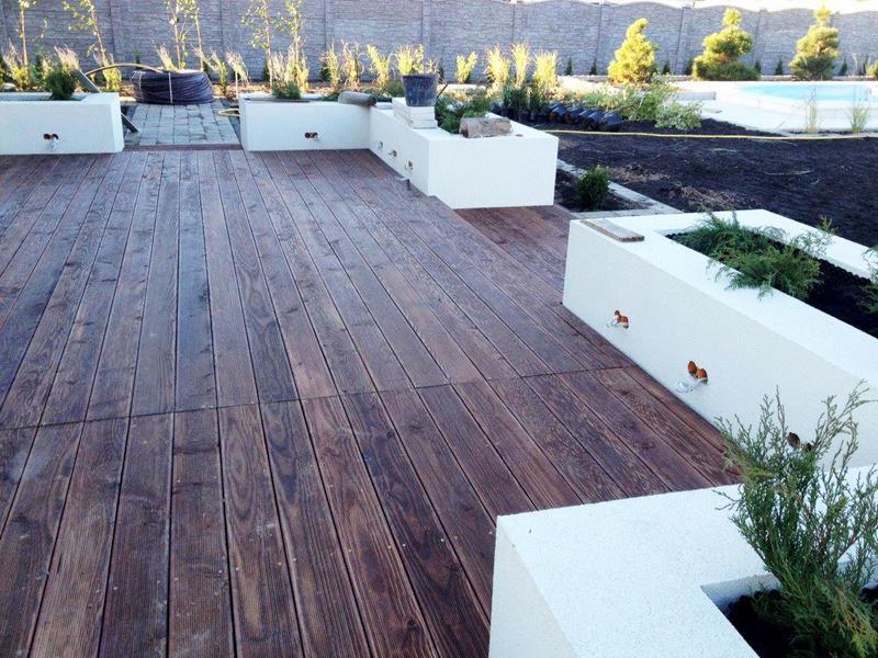 Ciemna, drewniana podłoga tarasu z białymi donicami, w tle mur i zieleń. Widoczne detale połączeń desek i faktura drewna. Minimalistyczny design.