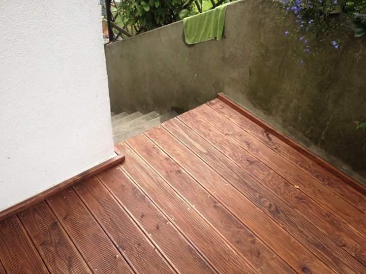 Drewniana podłoga tarasu z widocznymi deskami ułożonymi równolegle, obrzeża wykończone listwami. W tle ściana i schody.