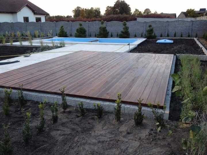 Drewniany taras z desek przy basenie, otoczony zielenią i ogrodzeniem z betonu. Ujęcie z perspektywy ogrodu, w tle dom jednorodzinny.