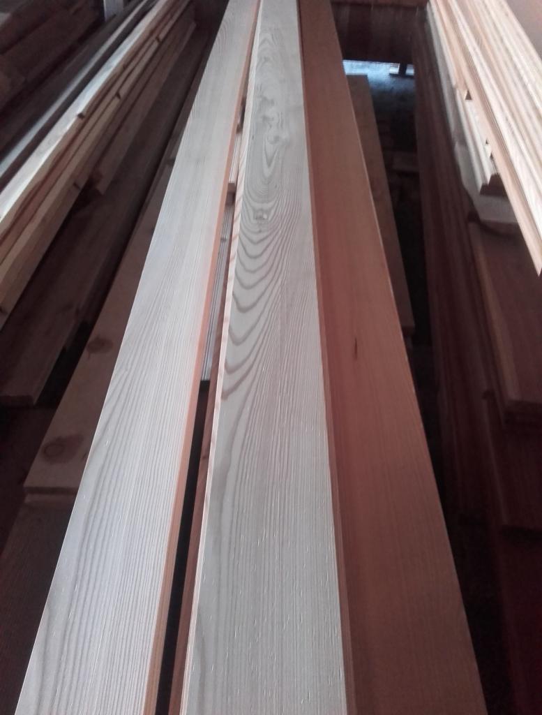 Jasne deski z wyraźnym rysunkiem słojów, ułożone w stos. Widoczne krawędzie i faktura drewna. Ujęcie z dołu podkreśla długość materiału.