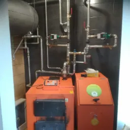 Pomarańczowy piec CO z podajnikiem, rury instalacji grzewczej na tle ciemnych płytek, zbiornik wyrównawczy u góry.