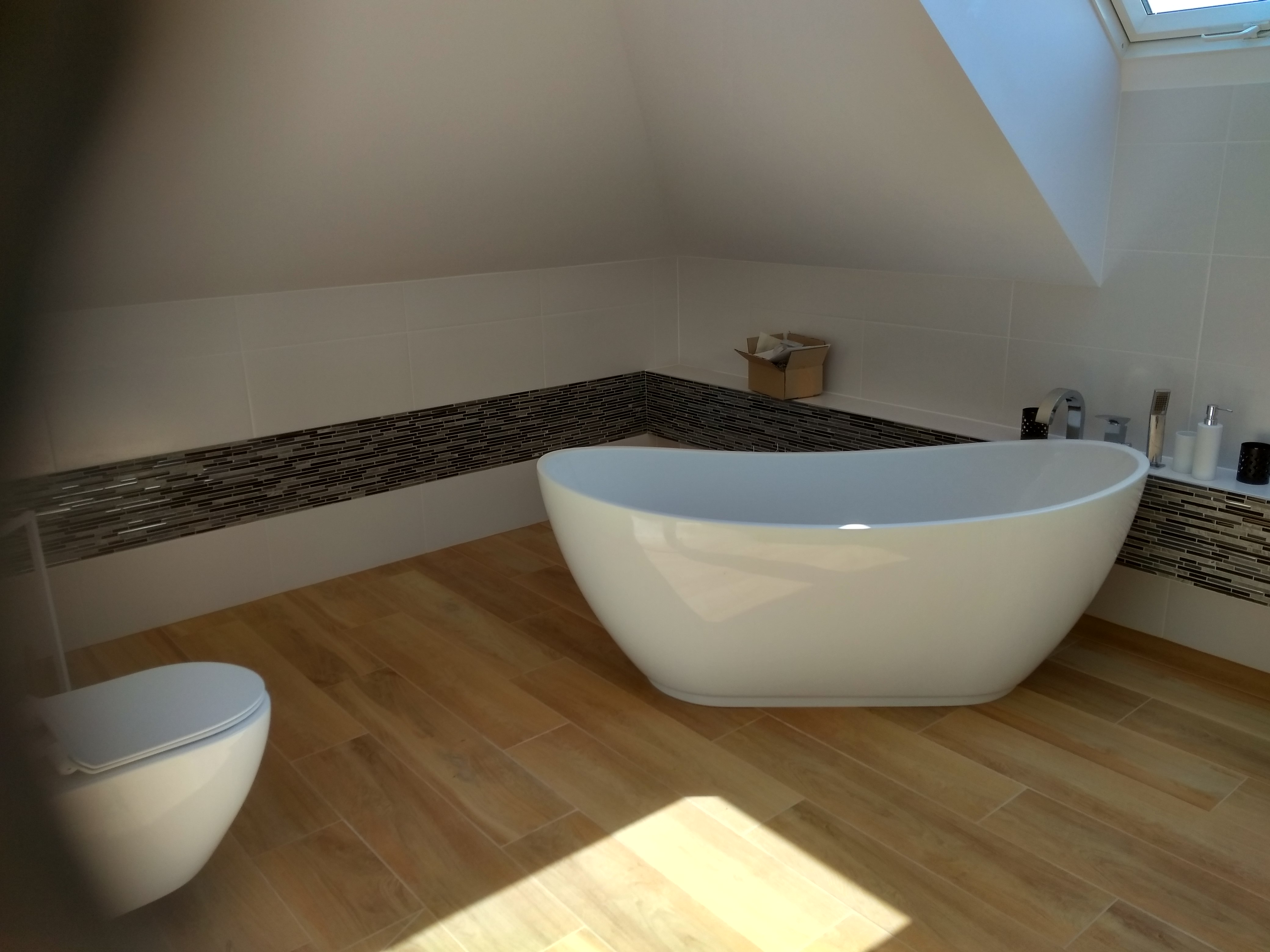 Nowoczesna łazienka na poddaszu z wolnostojącą białą wanną, wiszącą toaletą, drewnopodobną podłogą i mozaiką na ścianach.