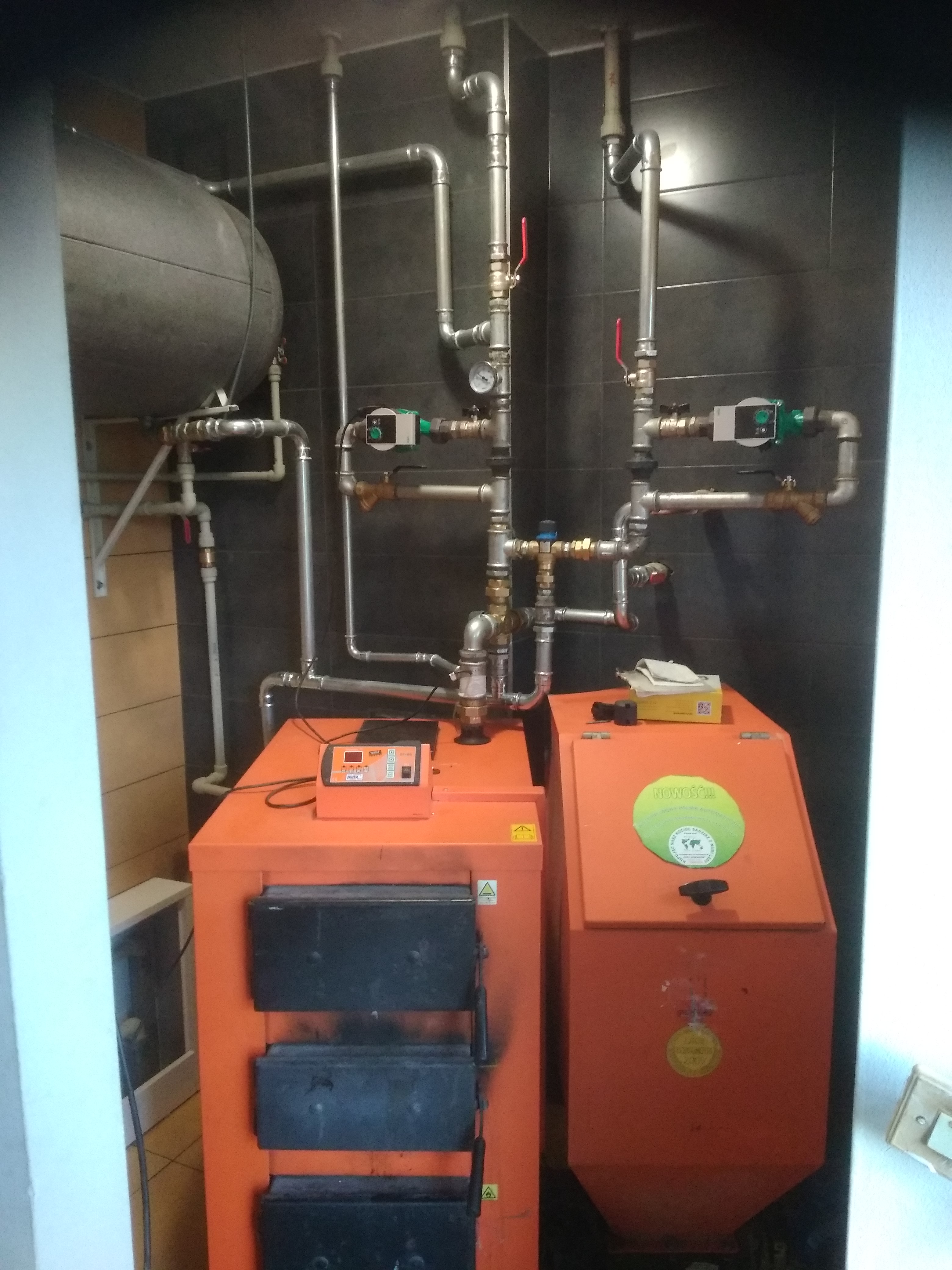 Pomarańczowy piec CO z podajnikiem, rury instalacji grzewczej na tle ciemnych płytek, zbiornik wyrównawczy u góry.