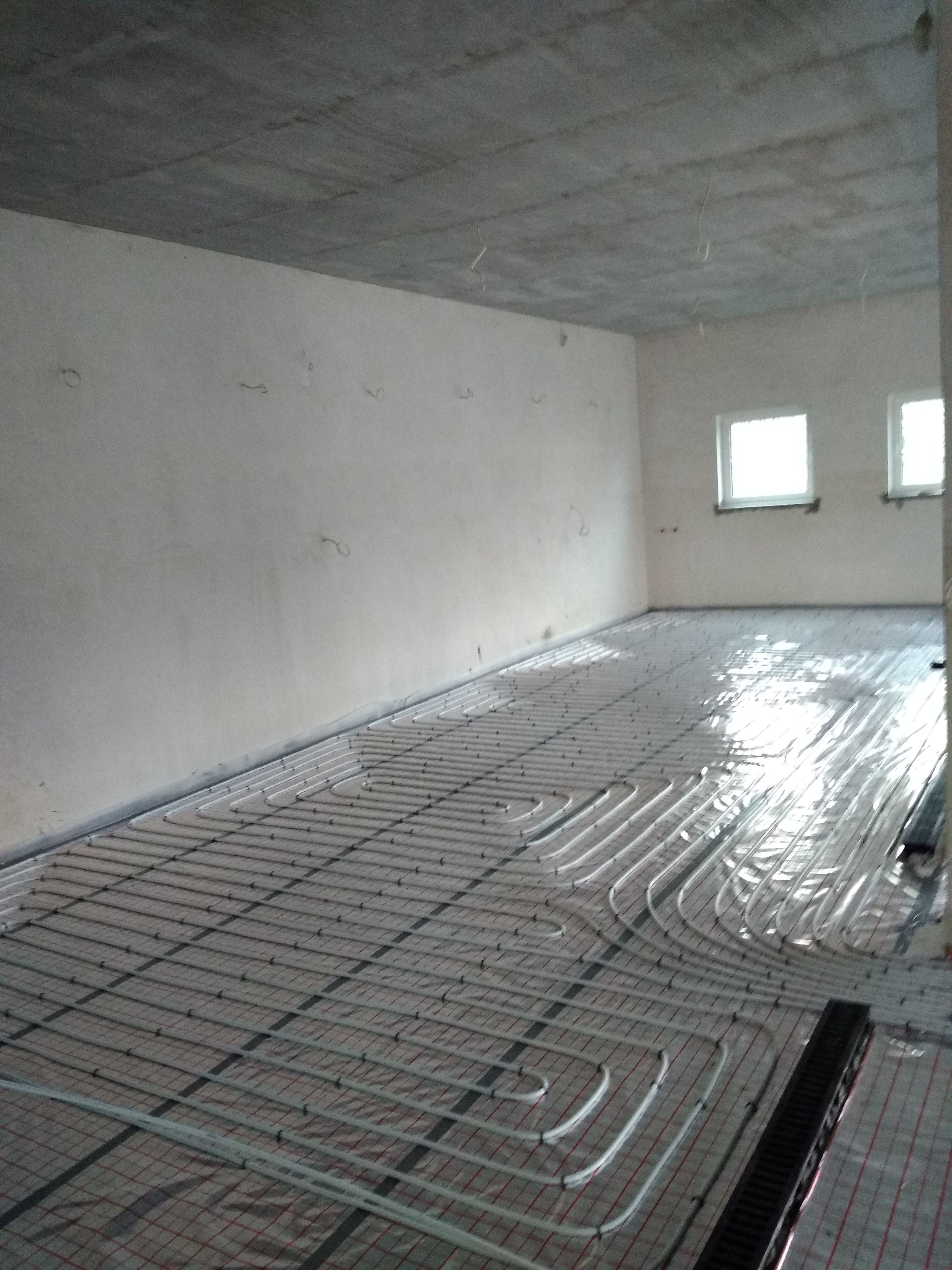 Białe rury ogrzewania podłogowego ułożone na folii izolacyjnej z rastrem, widok na surowy betonowy sufit i ściany w stanie deweloperskim, dwa okna w tle.