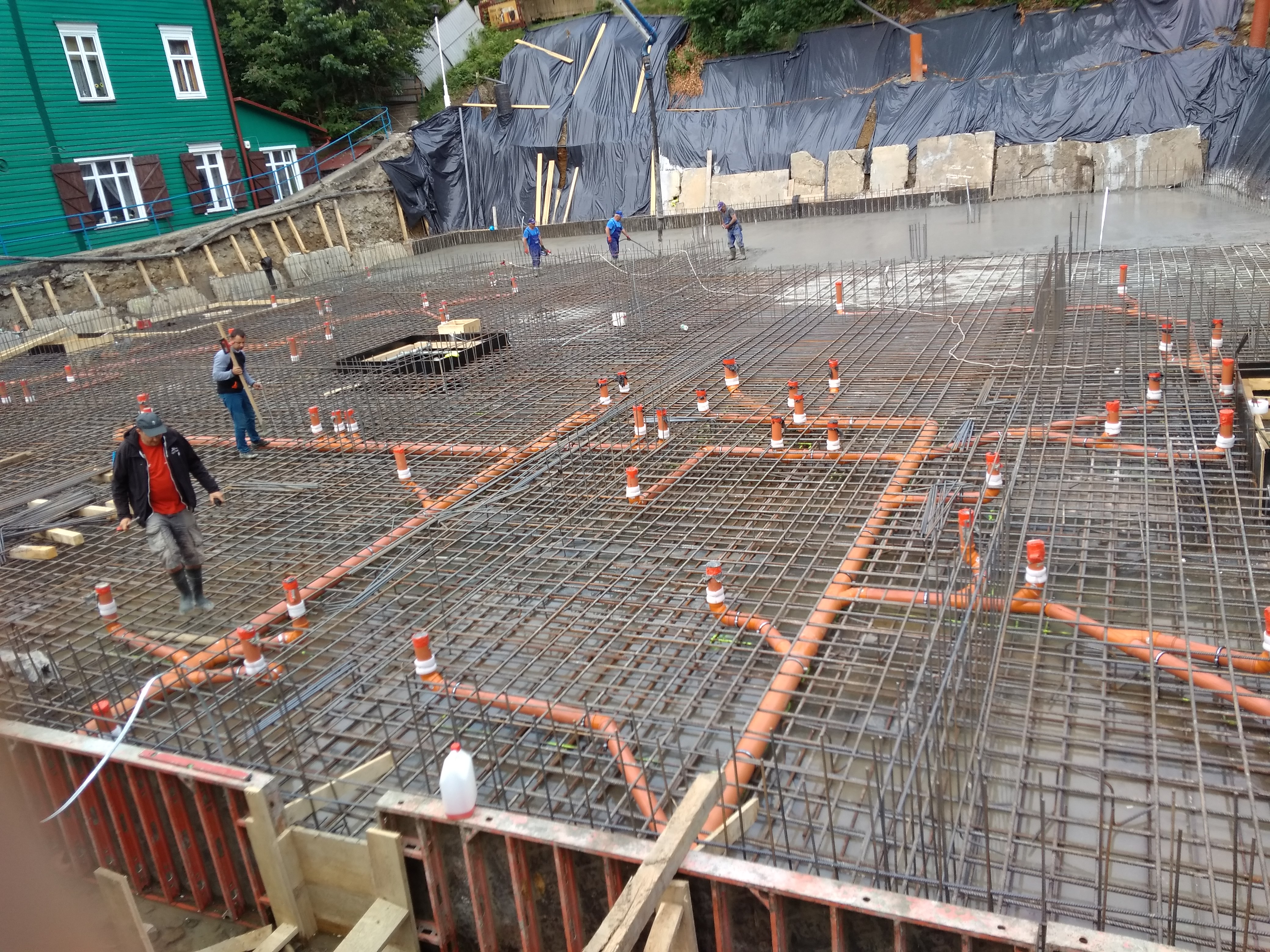 Siatka zbrojeniowa z wbudowanymi pomarańczowymi rurami kanalizacyjnymi na placu budowy, pracownicy w tle w trakcie wylewania betonu.