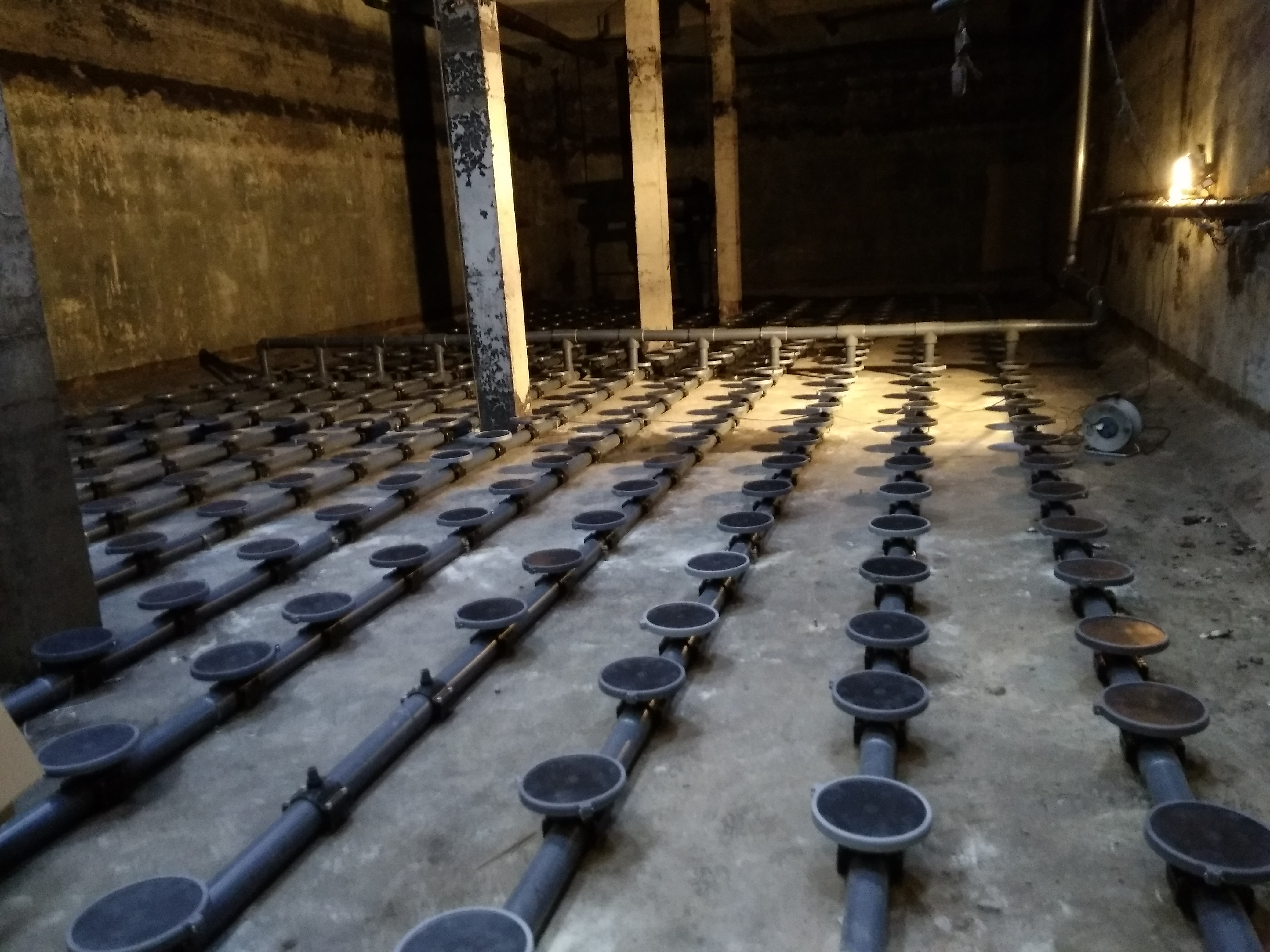 Instalacja dyfuzorów dyskowych w przydomowej oczyszczalni ścieków, widoczne rzędy szarych rur z czarnymi dyfuzorami na betonowym podłożu, w tle ściany zbiornika.