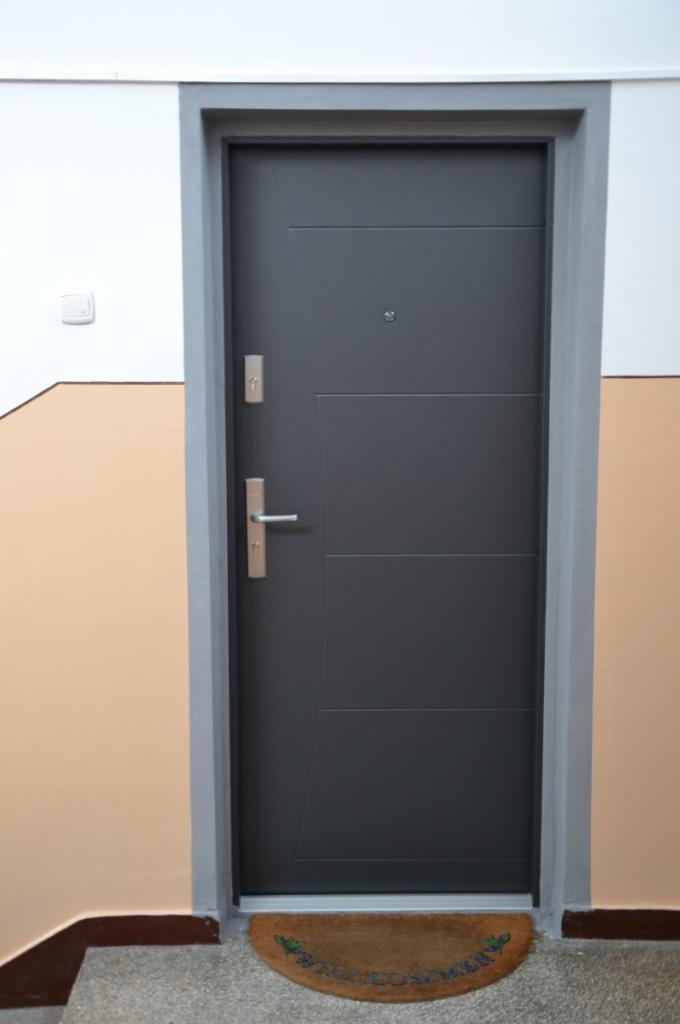 Szare drzwi zewnętrzne Wikęd z metalową ościeżnicą, widoczne od strony klatki schodowej z wycieraczką 'Welcome' przed nimi.