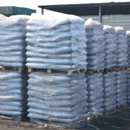 Rzędy palet z workami, szczelnie owiniętymi folią, składowane na zewnątrz, widoczne fragmenty etykiet na workach.