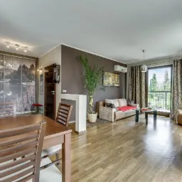 Jasne, przestronne wnętrze: drewniany stół z krzesłami, sofa, balkon z widokiem na zieleń. Na ścianie fototapeta z motywem górskiego jeziora. Nowoczesny wystrój.