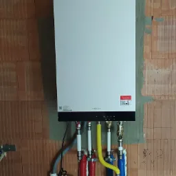 Nowoczesny, biały piec gazowy marki Viessmann zamontowany na ścianie z czerwonej cegły, z widocznymi kolorowymi rurami doprowadzającymi media i przewodami elektrycznymi.