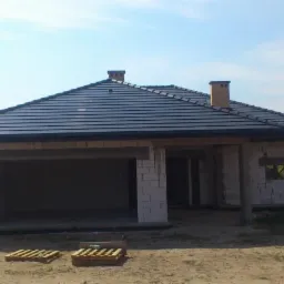 Dom w trakcie budowy z ciemną dachówką ceramiczną, widoczne kominy i elementy konstrukcyjne, palety na ziemi.