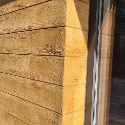 Elewacja domu z tynkiem imitującym drewno, widoczny narożnik budynku i okno z odbiciem lasu.