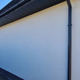 Świeżo pomalowana, jasna elewacja budynku z czarnymi rynnami i podbitką dachową w słoneczny dzień. Widoczny fragment elewacji sąsiedniego budynku i kontener na odpady budowlane w tle.