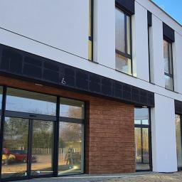 INTERBUD Mariusz Łyjak - Nowoczesny budynek biurowy z białymi ścianami, czarnymi akcentami i drewnianą okładziną, widoczne automatyczne drzwi wejściowe i odblaski na szybach.