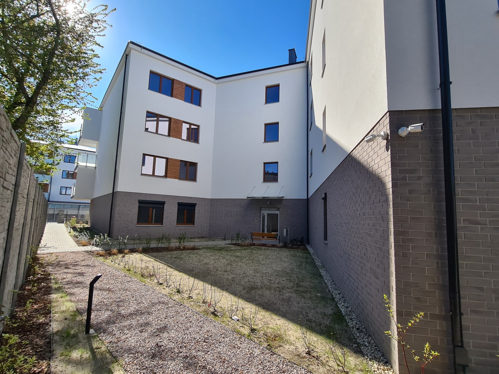 Nowoczesny budynek wielorodzinny z białą elewacją i szarym cokołem z cegły klinkierowej, widok z perspektywy ścieżki prowadzącej do wejścia, kamery monitoringu na ścianie.