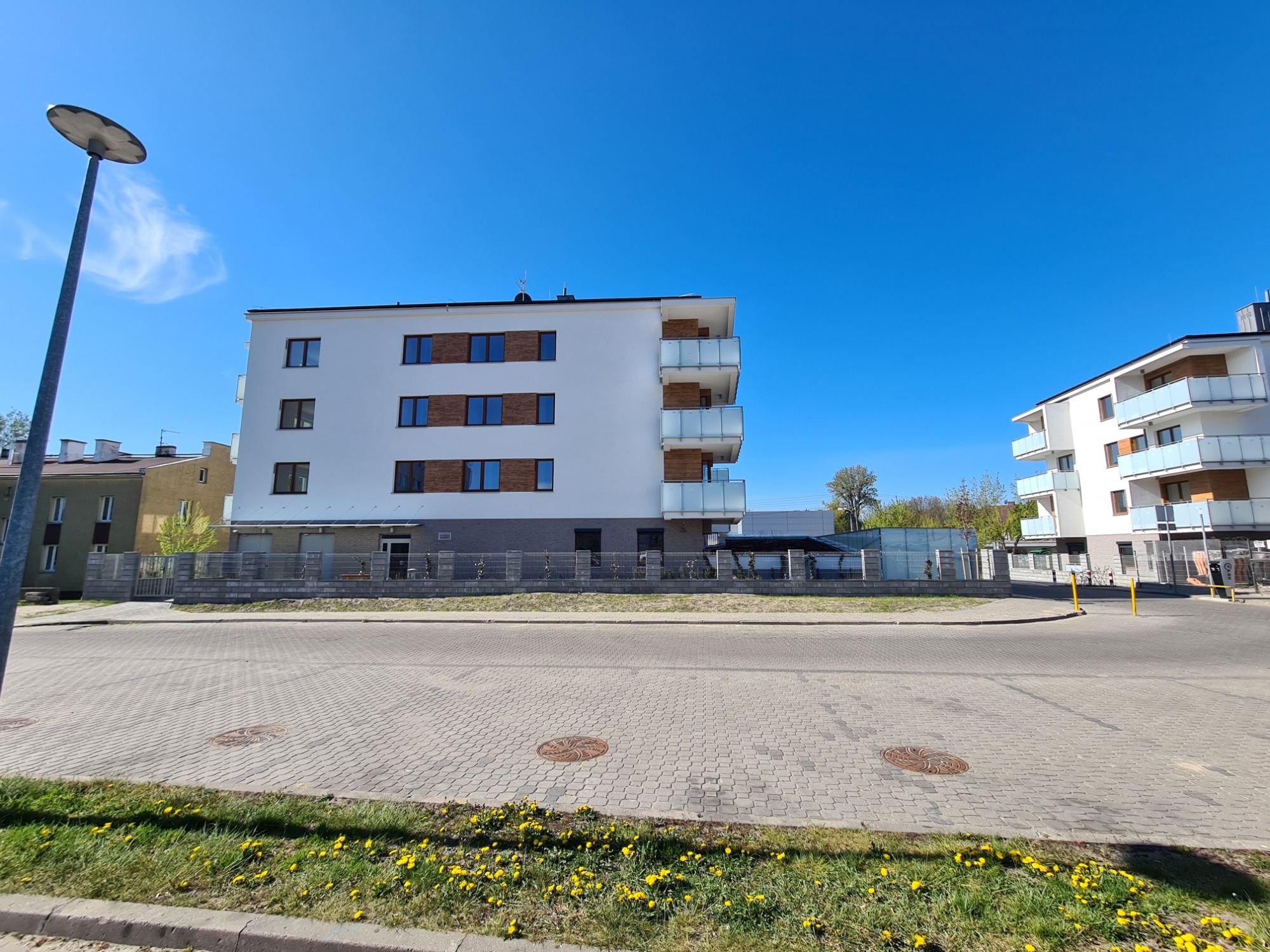 Nowoczesny budynek mieszkalny z białą elewacją i balkonami z przeszklonymi balustradami, widoczny z poziomu ulicy w słoneczny dzień. Dolna część elewacji wykończona szarą cegłą. W tle drugi budynek...