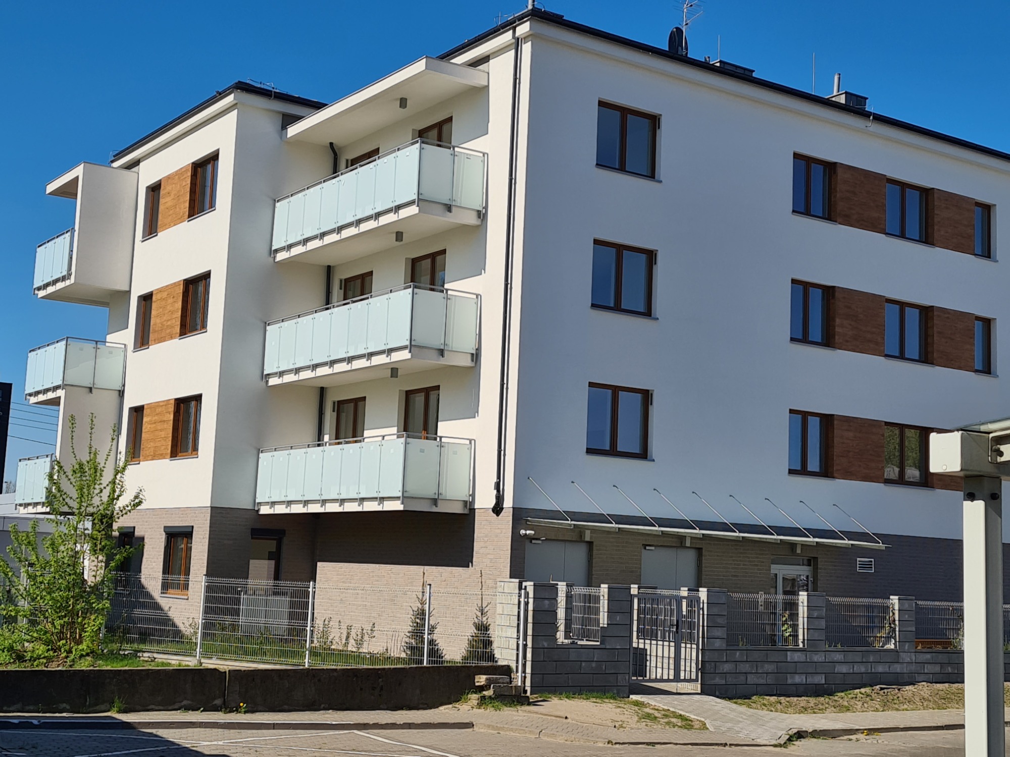 Nowoczesny budynek wielorodzinny z białymi elewacjami, balkonami z przezroczystymi balustradami i elementami dekoracyjnymi z drewnopodobnej okładziny, widziany z poziomu ulicy w słoneczny dzień.