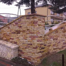 Kamienna ogrodowa konstrukcja z łukową, metalową poręczą, widziana z boku, w zimowej scenerii. Elementy z piaskowca, zróżnicowane odcienie.