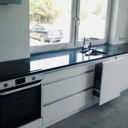 Kamieniarstwo Dziobak Radosław - Nowoczesna kuchnia z białymi szafkami i czarnym, błyszczącym blatem kamiennym pod oknem, widoczny fragment zlewu i bateria, na zewnątrz zaparkowany samochód.