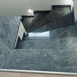 Kamieniarstwo Dziobak Radosław - Wewnętrzne schody z ciemnego kamienia z widoczną fakturą, oświetlone punktowym światłem sufitowym, z metalową listwą na krawędziach stopni.