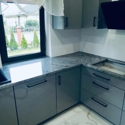 Kamieniarstwo Dziobak Radosław - Kuchenny blat z szarego kamienia z widocznym wzorem żyłkowania, zamontowany na szarych, lakierowanych szafkach z czarnymi uchwytami, z oknem w tle i widokiem na ogród.