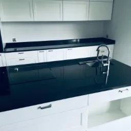 Błyszczący, czarny blat w nowoczesnej kuchni z białymi szafkami, widoczny zlew ze srebrną baterią i płyta indukcyjna.