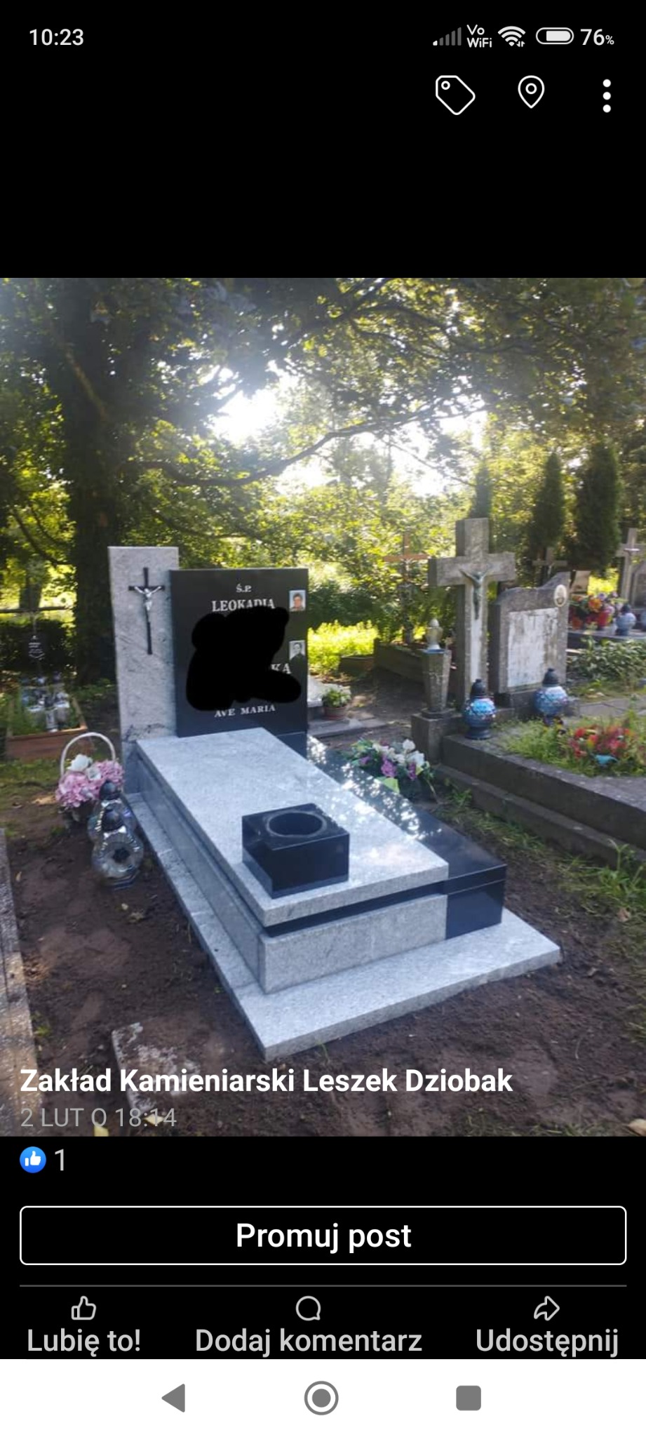 Szary nagrobek z czarną tablicą i krzyżem w słoneczny dzień na cmentarzu, widoczne kwiaty i znicze.