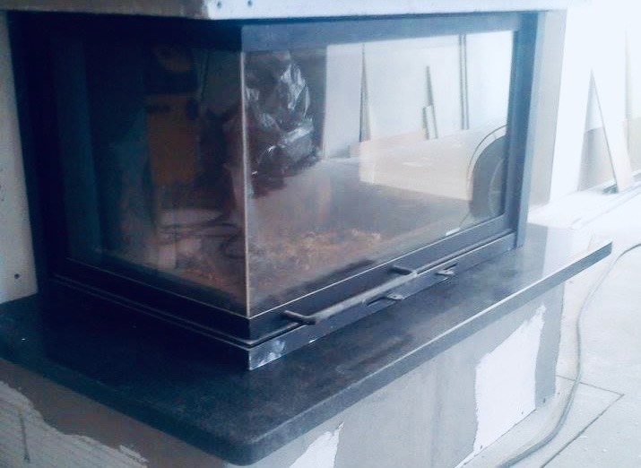 Ciemny, błyszczący blat pod kominkiem z czarną, metalową obudową i szklanymi drzwiczkami w trakcie montażu, widoczne niedoskonałości wykończenia ściany.