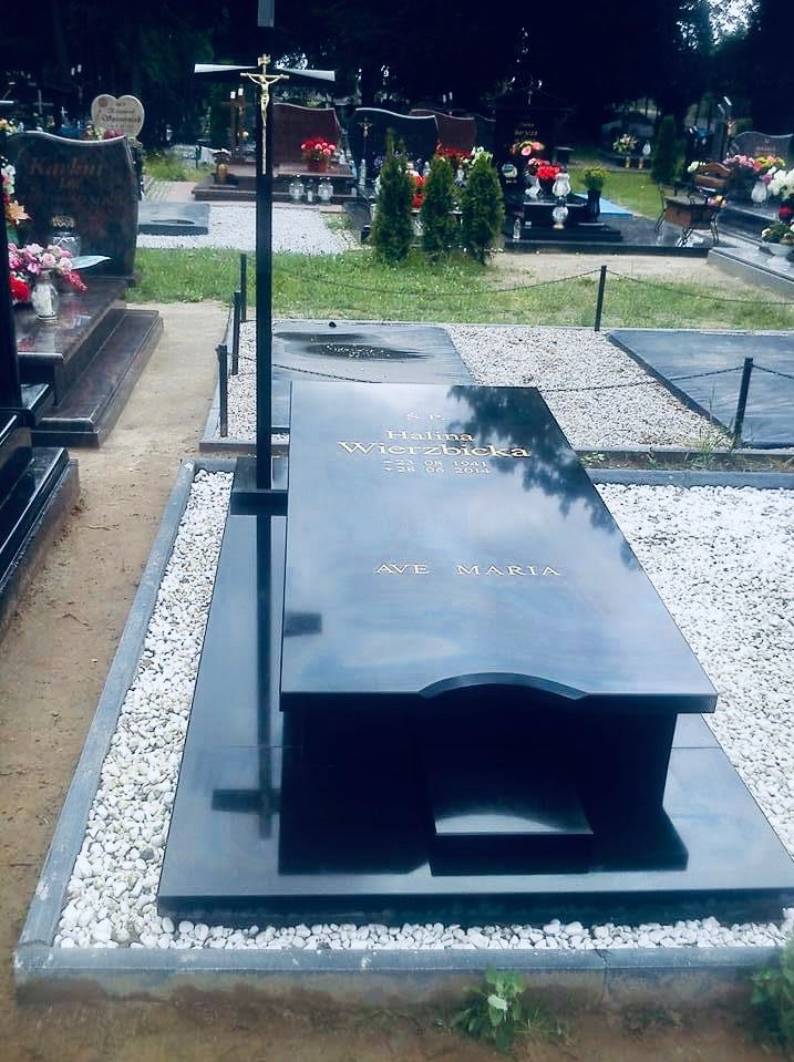 Czarny, polerowany nagrobek z inskrypcją 'Ave Maria' i metalowym krzyżem na cmentarzu, otoczony białym żwirem.