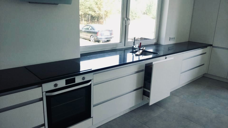 Nowoczesna kuchnia z białymi szafkami i czarnym, błyszczącym blatem kamiennym pod oknem, widoczny fragment zlewu i bateria, na zewnątrz zaparkowany samochód.