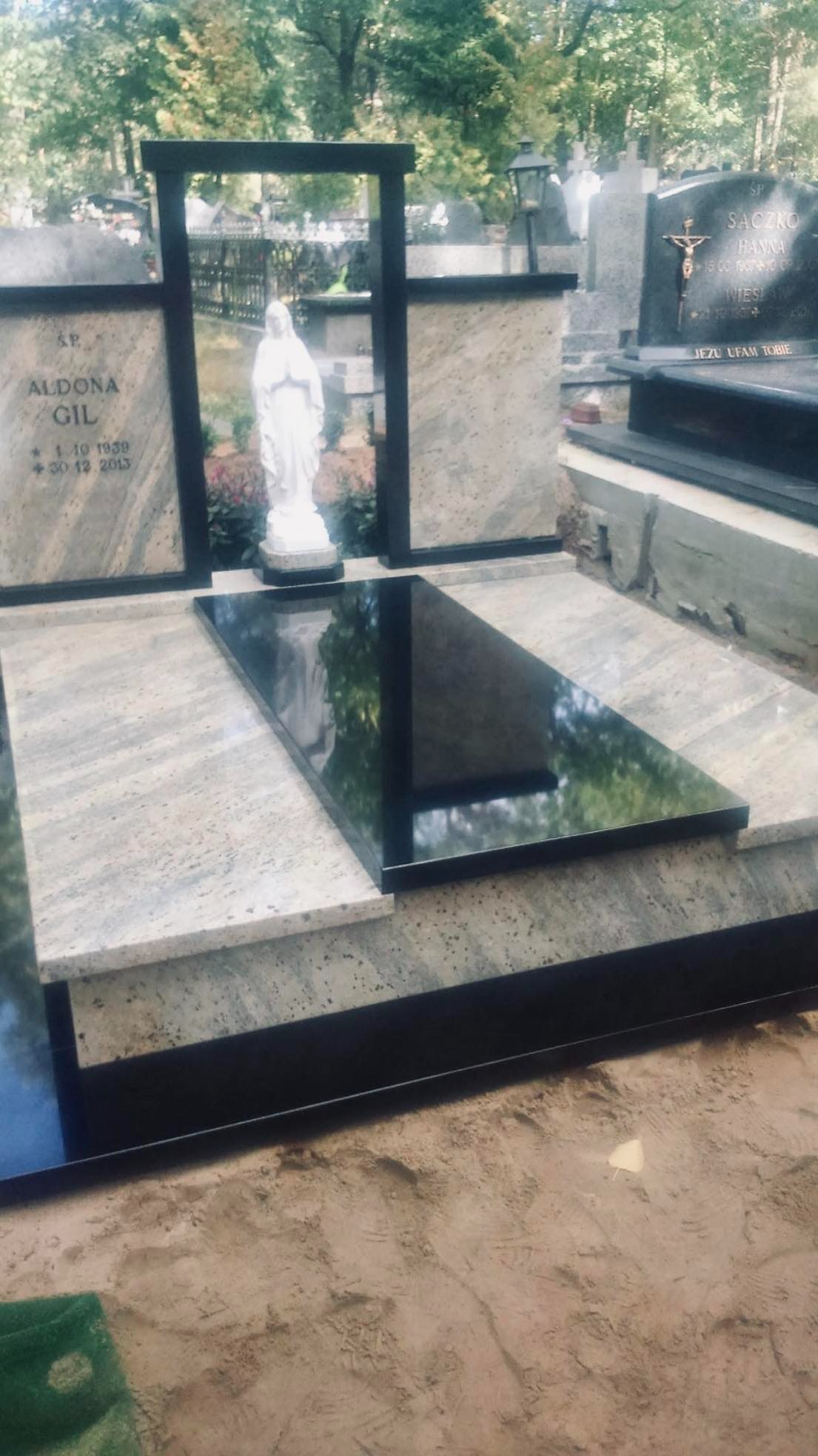 Nowoczesny nagrobek z jasnego granitu i czarnego kamienia, z rzeźbą Matki Boskiej w tle, na cmentarzu.