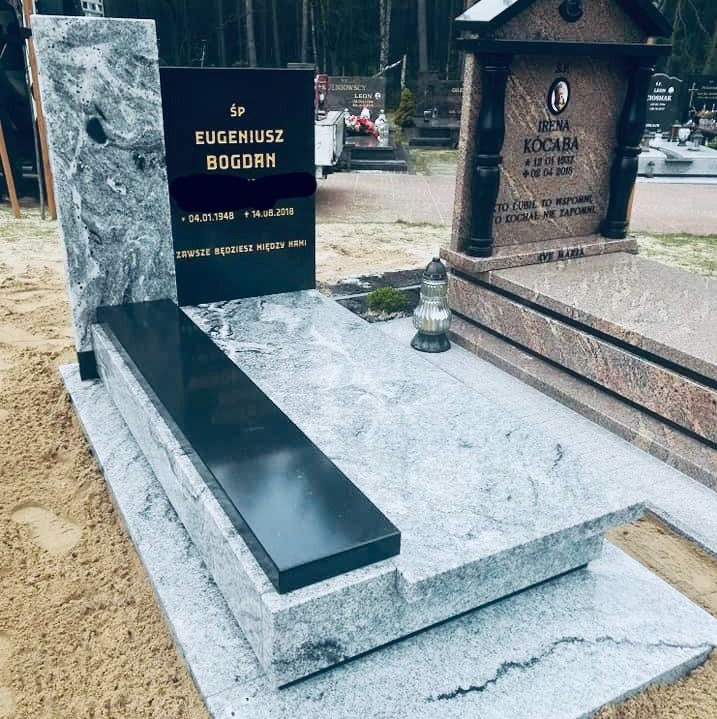 Granitowy nagrobek z czarną tablicą inskrypcyjną i elementami z szarego, wzorzystego kamienia, otoczony piaskiem na tle cmentarza z drzewami.