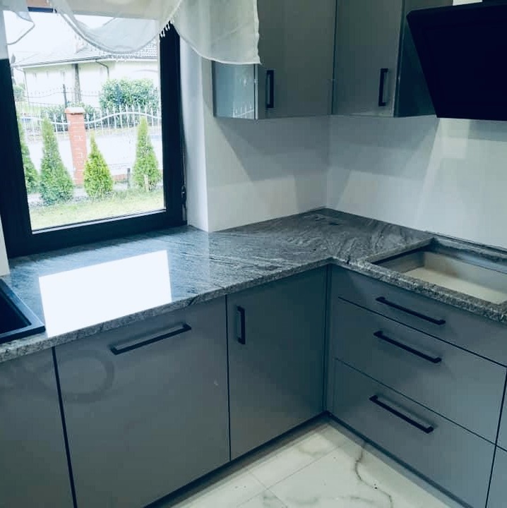 Kuchenny blat z szarego kamienia z widocznym wzorem żyłkowania, zamontowany na szarych, lakierowanych szafkach z czarnymi uchwytami, z oknem w tle i widokiem na ogród.