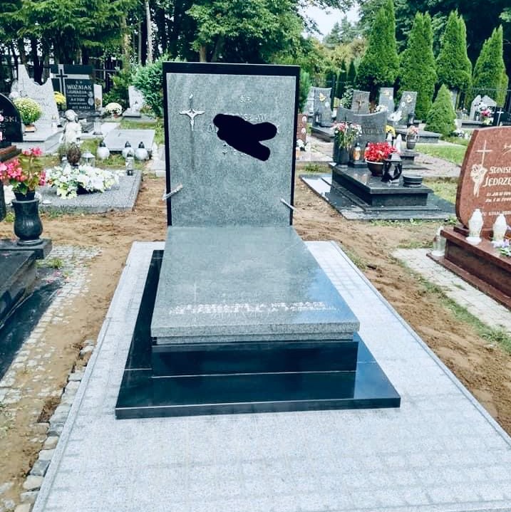 Nowoczesny, szary nagrobek z czarną podstawą na cmentarzu, z widocznymi innymi nagrobkami w tle.