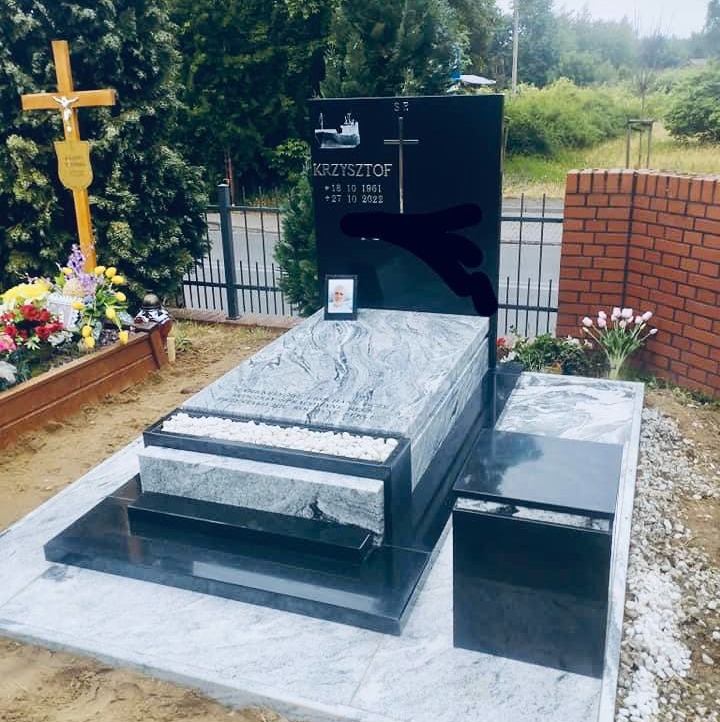 Czarny nagrobek z szarą płytą wierzchnią, ozdobiony fotografią zmarłego i krzyżem, otoczony kwiatami i ceglanym murem.