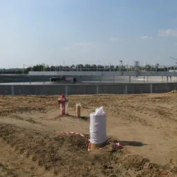 Budowa oczyszczalni ścieków: widok na betonowy zbiornik z wodą, hydrant i zabezpieczone rury na piaszczystym terenie w słoneczny dzień.