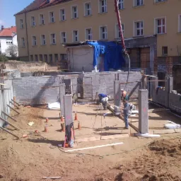 Fundamenty budynku w trakcie budowy: betonowe słupy, ściany z bloczków, zbrojenia. Robotnicy przy pracy, widoczna pompa do betonu. Ujęcie z góry, w tle budynek.